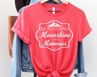 Könnte beinhalten: Rotes T-Shirt mit einem weißen Grafikdesign, auf dem in einer Vintage-Schriftart "Mountains Moonshine and Memories" steht.