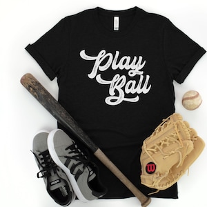 Peut inclure: Un t-shirt noir avec le texte "Play Ball" en lettres blanches vieillies. Le t-shirt est pr&eacute;sent&eacute; avec une batte de baseball, une balle de baseball et un gant de baseball.