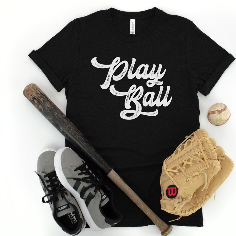 Peut inclure: Un t-shirt noir avec le texte "Play Ball" en lettres blanches vieillies. Le t-shirt est pr&eacute;sent&eacute; avec une batte de baseball, une balle de baseball et un gant de baseball.