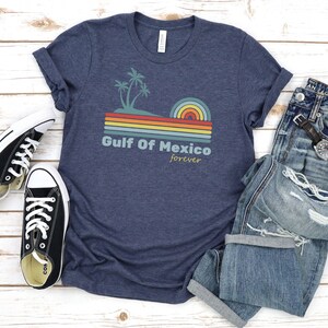 Pu&ograve; includere: Una maglietta blu navy con una grafica retr&ograve; di palme, un tramonto e il testo "Golfo del Messico per sempre".