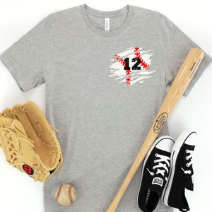 K&ouml;nnte beinhalten: Graues T-Shirt mit der Nummer 12 in einem schwarzen und roten Baseball-Naht-Design. Ein Baseballhandschuh, ein Schl&auml;ger, ein Baseball und schwarze Converse-Sneaker sind um das Shirt angeordnet.