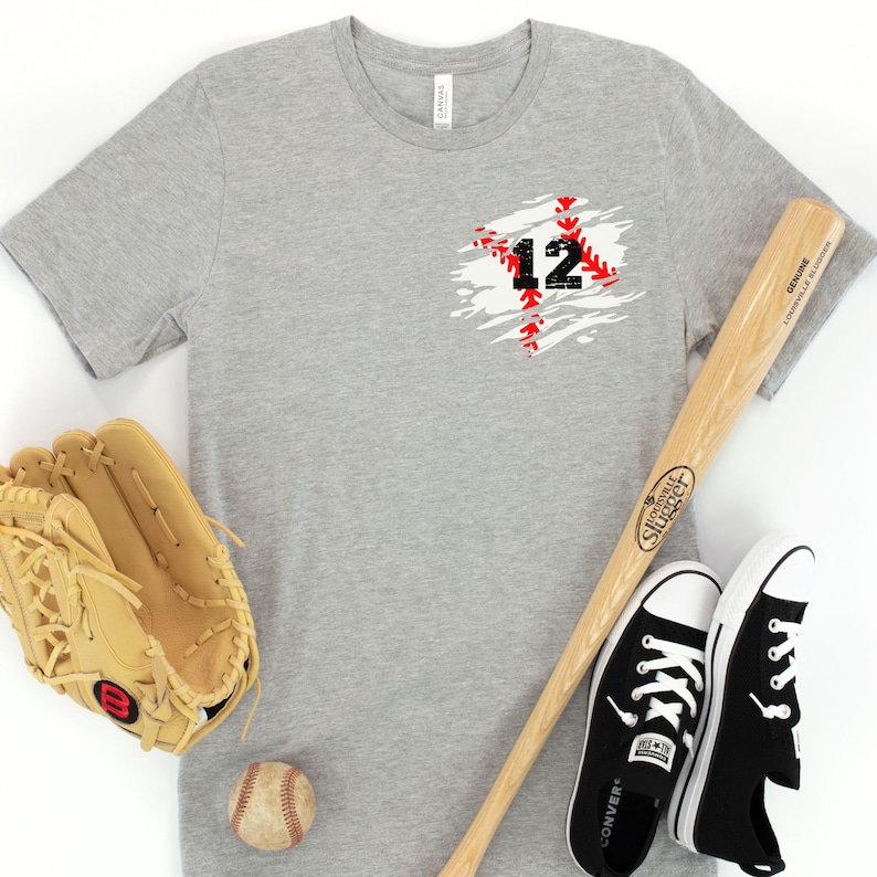 K&ouml;nnte beinhalten: Graues T-Shirt mit der Nummer 12 in einem schwarzen und roten Baseball-Naht-Design. Ein Baseballhandschuh, ein Schl&auml;ger, ein Baseball und schwarze Converse-Sneaker sind um das Shirt angeordnet.