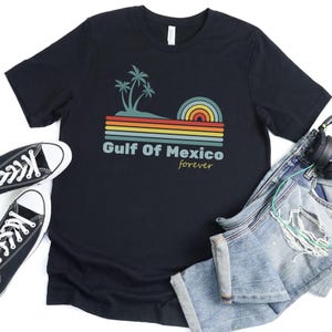Pu&ograve; includere: Maglietta nera con una grafica retr&ograve; di palme e un tramonto su uno sfondo a righe. Il testo "Gulf of Mexico forever" &egrave; stampato sotto la grafica.