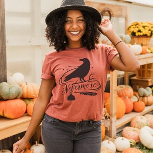 May include: A rust-colored t-shirt with a black raven silhouette and the text "Nevermore" in a stylized font. The raven is perched on a branch with a quill and inkwell below.