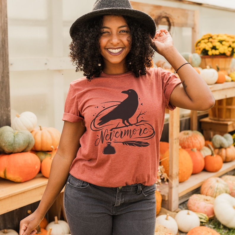 May include: A rust-colored t-shirt with a black raven silhouette and the text "Nevermore" in a stylized font. The raven is perched on a branch with a quill and inkwell below.