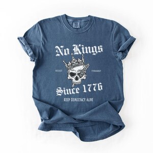 Może przedstawiać: Niebieska koszulka z białym napisem "No Kings", czaszką w koronie i słowami "Since 1776". Koszulka zawiera r&oacute;wnież frazę "Keep Democracy Alive". Rękawy są podwinięte.