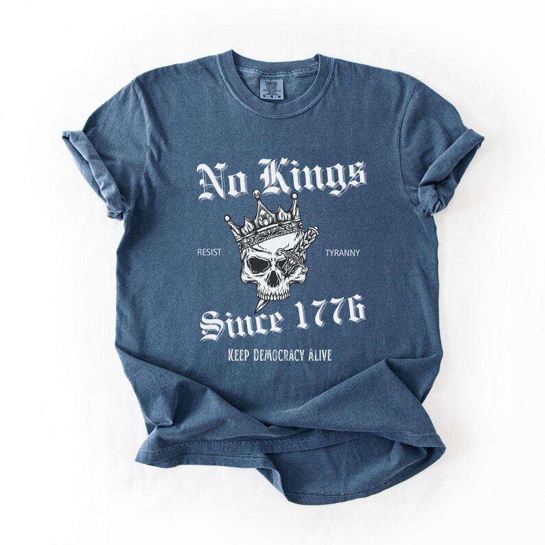 Może przedstawiać: Niebieska koszulka z białym napisem "No Kings", czaszką w koronie i słowami "Since 1776". Koszulka zawiera r&oacute;wnież frazę "Keep Democracy Alive". Rękawy są podwinięte.