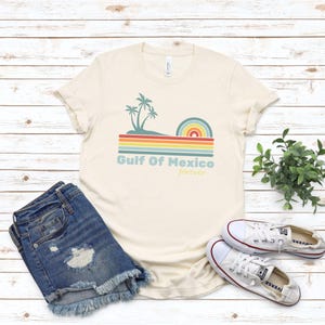 Pu&ograve; includere: Una maglietta beige con un design grafico retr&ograve; che presenta palme, un arcobaleno e il testo "Golfo del Messico per sempre".
