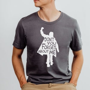 Puede incluir: Una camiseta gris oscuro con una silueta blanca de una persona con el texto "Don't you forget about me" impreso en la parte delantera.