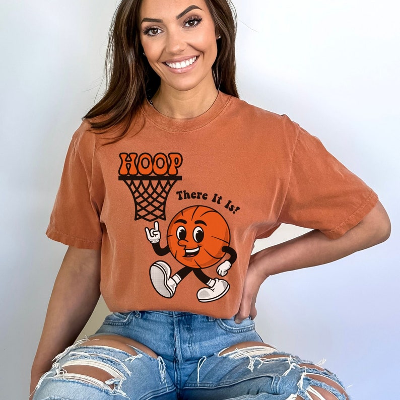 May include: A burnt orange t-shirt with a cartoon basketball character and the text "HOOP There It Is!" printed on the front.