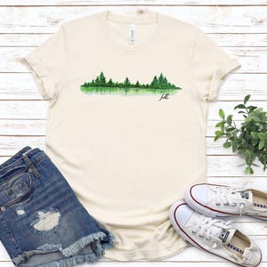 May include: A cream colored t-shirt with a watercolor design of a forest reflected in a lake. The text "felt" is written below the design.