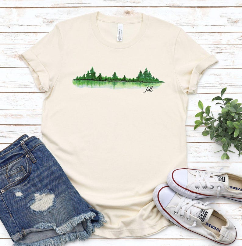 May include: A cream colored t-shirt with a watercolor design of a forest reflected in a lake. The text "felt" is written below the design.