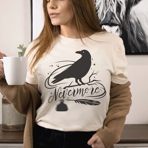 May include: A white t-shirt with a black raven illustration and the text "Nevermore" in a stylized font. The raven is perched on a branch with a quill pen and inkwell below.