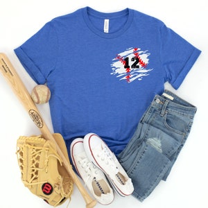K&ouml;nnte beinhalten: Ein blaues T-Shirt mit Baseball-Design, das die Nummer 12 in Schwarz und Rot zeigt. Das Bild enth&auml;lt einen Baseball, einen Schl&auml;ger, einen Handschuh, Jeans und wei&szlig;e Turnschuhe, was eine sportliche &Auml;sthetik erzeugt.