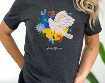 May include: Dark gray t-shirt featuring a white dove carrying a sprig, set against a Ukrainian flag design. The design includes blue and yellow flowers and the text "Slava Ukraini".