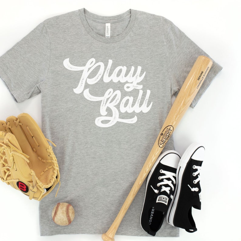 Peut inclure: Un t-shirt gris avec les mots "Play Ball" en &eacute;criture blanche. Une batte de baseball, un gant de baseball, une balle de baseball et des baskets noires sont dispos&eacute;s autour du t-shirt.