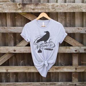 May include: A gray t-shirt with a black raven silhouette and the text "Nevermore" printed on the front. The raven is perched on a branch with a quill pen and inkwell below.
