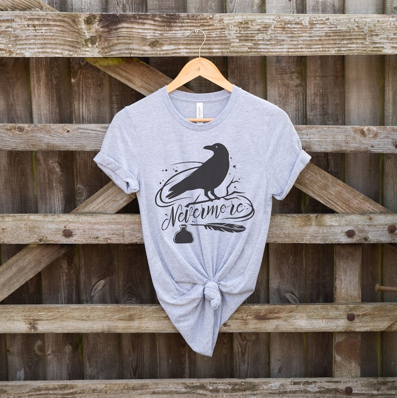 May include: A gray t-shirt with a black raven silhouette and the text "Nevermore" printed on the front. The raven is perched on a branch with a quill pen and inkwell below.