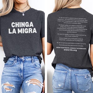 May include: Dark gray t-shirt with the text "CHINGA LA MIGRA" in white. The back of the shirt has text in English and Spanish about constitutional rights. The shirt is paired with blue jeans.