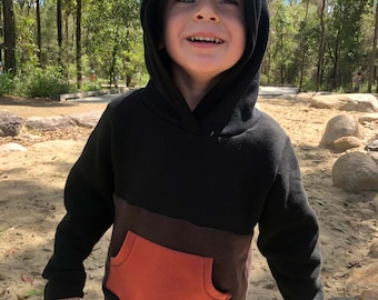 May include: A black, brown and orange colour block hoodie with a kangaroo pocket. The hoodie has a drawstring hood and is being worn by a child.