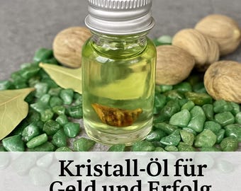 May include: A small glass bottle with a silver lid filled with a green liquid and a small piece of amber-colored crystal. The bottle is surrounded by green and brown stones. The text on the label reads "Kristall-öl für Geld und Erfolg Crystal Money and Success Oil".