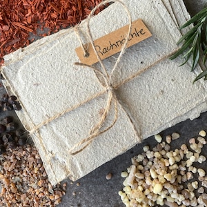 Könnte beinhalten: Ein Stapel handgemachtes Papier, mit Schnur gebunden, mit einem kleinen Etikett mit der Aufschrift "Rauchnächte". Das Papier ist von verschiedenen natürlichen Elementen umgeben, darunter rötlich-braune Holzspäne, kleine schwarze Pfefferkörner und helles Harz.