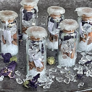 Può includere: Sei piccole bottiglie di vetro riempite con cristalli bianchi, fiori secchi e bastoncini di cannella. Le bottiglie sono sormontate da un sigillo di cera bianca e tappi di sughero.