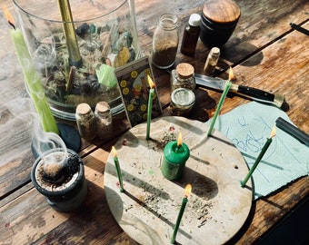 May include: A wooden table with a tarot card, a glass jar, candles, and other items used in a spiritual ritual. The tarot card is the Star card, which is associated with hope, inspiration, and renewal. The candles are green and are burning. There is a small green candle in the center of the table, surrounded by a circle of green candles. The candles are arranged in a circle around a small green candle in the center. There is a piece of paper with writing on it, and a small bowl with incense burning.