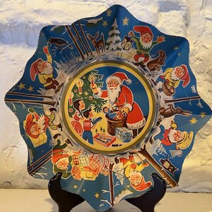 May include: A blue and white paper plate with a scalloped edge features a Christmas scene with Santa Claus, elves, and a Christmas tree. The plate is decorated with stars and other festive details.