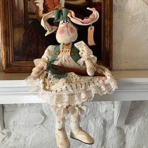Könnte beinhalten: Eine handgefertigte Stoffhasenpuppe, die eine grüne Weste und ein weißes Kleid mit einer Spitzenborte trägt. Die Puppe hat eine rosa Nase und grüne Augen. Die Puppe sitzt auf einem weißen Kaminsims mit einem Gemälde im Hintergrund.