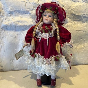 May include: A porcelain doll with blonde hair and blue eyes wearing a red velvet dress with white lace trim and a red hat with roses. The doll is holding a white purse.