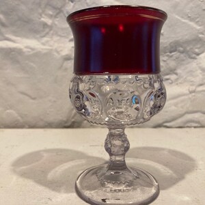 Puede incluir: Una copa de vidrio transparente con un borde rojo y un patrón texturizado. La copa tiene una base redonda y un tallo.