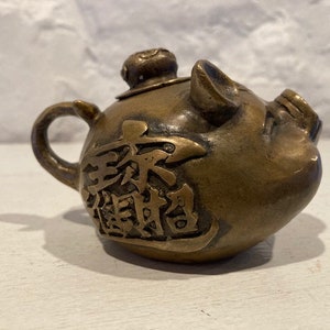 May include: A small, bronze teapot shaped like a pig. The teapot has Chinese characters on the side and a lid with a small knob.