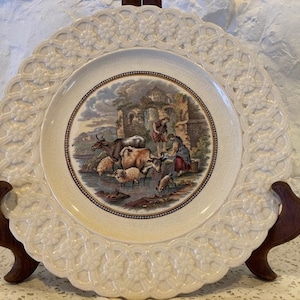 Könnte beinhalten: Ein weißer Keramikteller mit einem erhabenen, komplizierten Design am Rand. Die Mitte des Tellers zeigt eine ländliche Szene mit einem Bauernhaus, Tieren und Figuren. Die Szene ist in gedämpften Tönen von Braun, Grün und Blau gemalt.