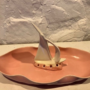 May include: A pink ceramic dish with a white ceramic sailboat figurine in the centre. The dish has a scalloped edge and the sailboat has a hole in the bottom for holding toothpicks.