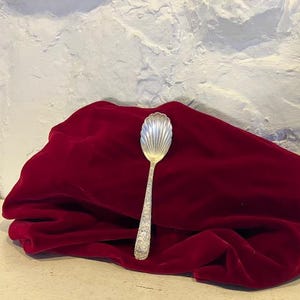 Puede incluir: Una cuchara plateada con un cuenco en forma de concha y un mango adornado descansa sobre una tela de terciopelo rojo oscuro. El cuenco de la cuchara tiene un diseño acanalado y el mango presenta intrincados patrones florales. El fondo es una pared blanca texturizada.