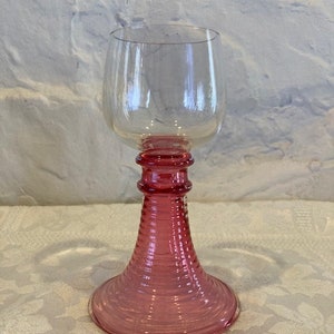 May include: A clear glass wine glass with a pink, ribbed stem and base. The glass has a round bowl and a wide base.