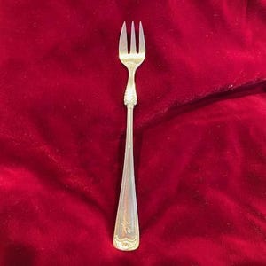 Op de afbeelding: Een zilveren vork met drie tanden, rustend op een dieprode fluwelen achtergrond. De vork heeft een decoratief handvat met sierlijke details. De tanden zijn gelijkmatig verdeeld en de steel loopt taps toe.