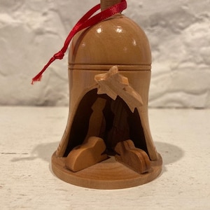 May include: A wooden Christmas bauble shaped like a bell with a red ribbon. The bell has a carved nativity scene inside, depicting Mary, Joseph, and the baby Jesus.