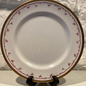 May include: A white porcelain dinner plate with a gold rim and a delicate pink floral pattern. The plate is on a stand.