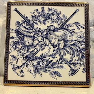 Puede incluir: Azulejo de cerámica cuadrado con fondo crema y diseño floral y cinta azul y blanco. Dos instrumentos cruzados en forma de trompeta están en el centro. El azulejo tiene un borde dorado.