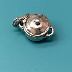 May include: A small, three-dimensional silver-toned charm shaped like a miniature cooking pot with a lid and two side handles. The charm features a rounded body, a domed lid with a central knob, and a small circular jump ring attached to the side.