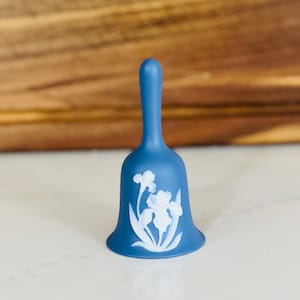 May include: A blue ceramic bell with a white floral design. The bell has a long handle and a flared base. The floral design features white iris flowers. The bell is likely a decorative item.