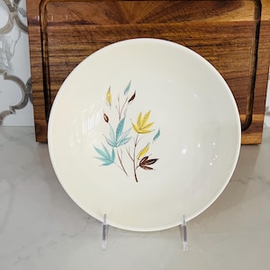 May include: A white ceramic bowl with a floral design featuring blue, yellow, and brown leaves. The bowl is on a wooden cutting board.