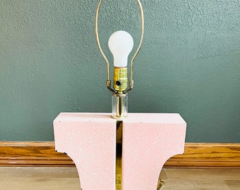 May include: A pink and gold table lamp with a clear glass shade and a white light bulb. The lamp base is made of a textured material and has a gold accent.