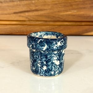 May include: A small, cylindrical ceramic vessel featuring a mottled, deep blue and white marbled glaze pattern. The container has a thick, rounded rim and a smooth, glossy finish, with a white interior visible at the top opening.