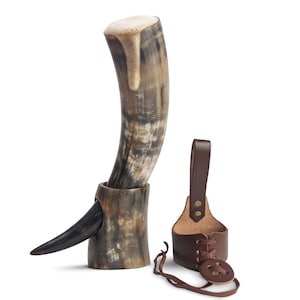 May include: A curved drinking horn with a natural mottled brown and cream finish, resting in a matching horn stand with a pointed side protrusion. Beside it sits a brown leather belt holster featuring a lace-up design and a vertical strap.