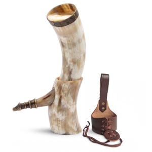 May include: A curved drinking horn with a natural cream and brown mottled finish, featuring a metallic rim with intricate embossed patterns and a pointed metal tip. Beside it sits a brown leather holster with a lace-up design and a belt loop.