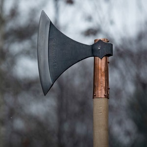 May include: A black axe with a copper handle and the word "SKARTH" inscribed on the blade. The axe is set against a blurred background of trees.