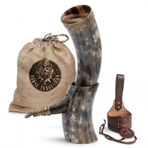May include: A drinking horn with a polished, mottled grey and black finish, featuring a brass rim with an etched knotwork pattern. It includes a brown leather belt holster with lace-up details and a tan burlap drawstring bag labeled Norse Tradesman.
